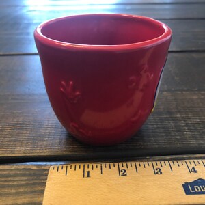 May include: A red ceramic mug with a white and blue design. The mug is approximately 3 inches tall.