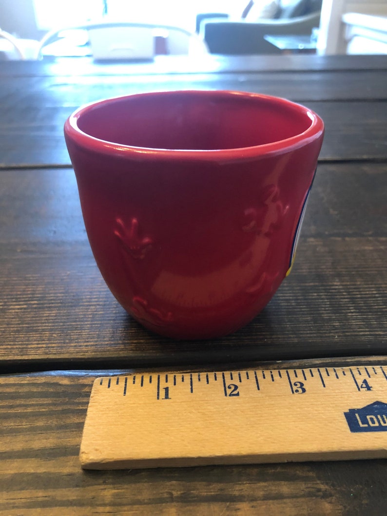May include: A red ceramic mug with a white and blue design. The mug is approximately 3 inches tall.