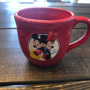May include: Red ceramic mug with a white circle featuring Mickey Mouse and Minnie Mouse holding hands. The mug has a white handle and a white star design.