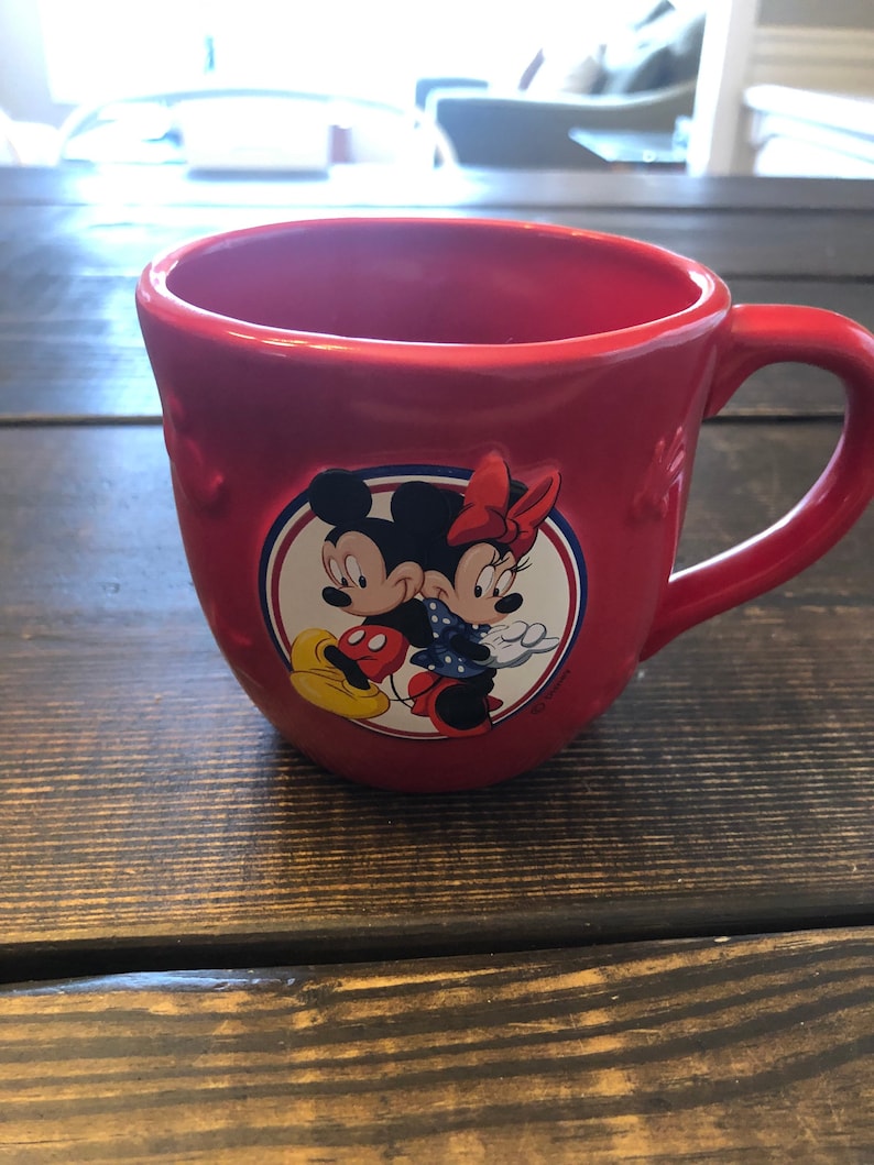 May include: Red ceramic mug with a white circle featuring Mickey Mouse and Minnie Mouse holding hands. The mug has a white handle and a white star design.