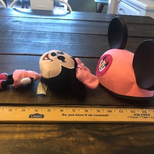 May include: A pink Minnie Mouse hat with black ears and a red Minnie Mouse logo. A plush Minnie Mouse toy is lying on a wooden surface.
