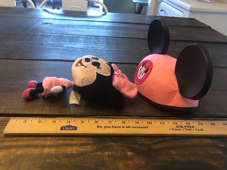 May include: A pink Minnie Mouse hat with black ears and a red Minnie Mouse logo. A plush Minnie Mouse toy is lying on a wooden surface.