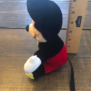 May include: A black and red plush Mickey Mouse toy with a white face and yellow shoes. The toy is sitting on a wooden surface and has a long black tail.