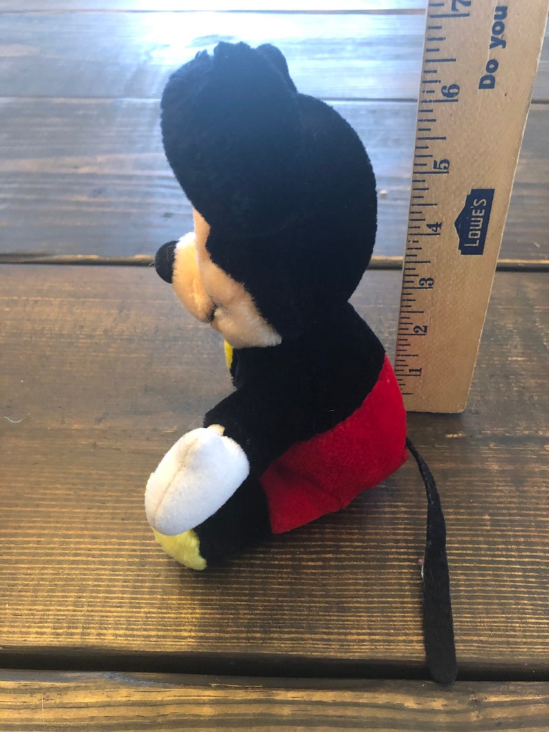 May include: A black and red plush Mickey Mouse toy with a white face and yellow shoes. The toy is sitting on a wooden surface and has a long black tail.
