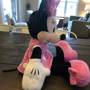 May include: A plush pink and black Minnie Mouse toy with white polka dots on her dress and a pink bow on her head. She is sitting with her legs crossed and her right arm outstretched.