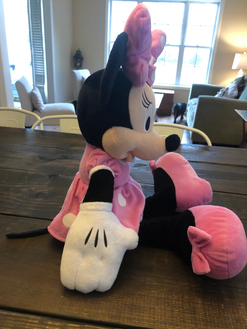 May include: A plush pink and black Minnie Mouse toy with white polka dots on her dress and a pink bow on her head. She is sitting with her legs crossed and her right arm outstretched.