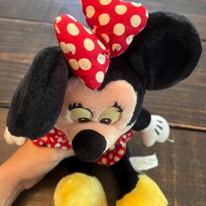 May include: A plush Minnie Mouse toy with a red and white polka dot bow, black ears, and yellow shoes. The toy is sitting on a wooden surface.