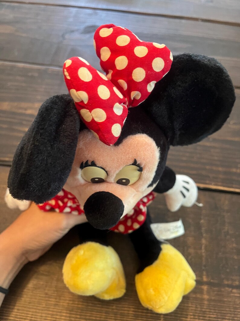 May include: A plush Minnie Mouse toy with a red and white polka dot bow, black ears, and yellow shoes. The toy is sitting on a wooden surface.