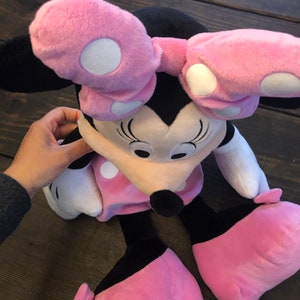 May include: Pink and black plush toy of Minnie Mouse with white polka dots. The toy has a large pink bow and is sitting on a wooden surface.
