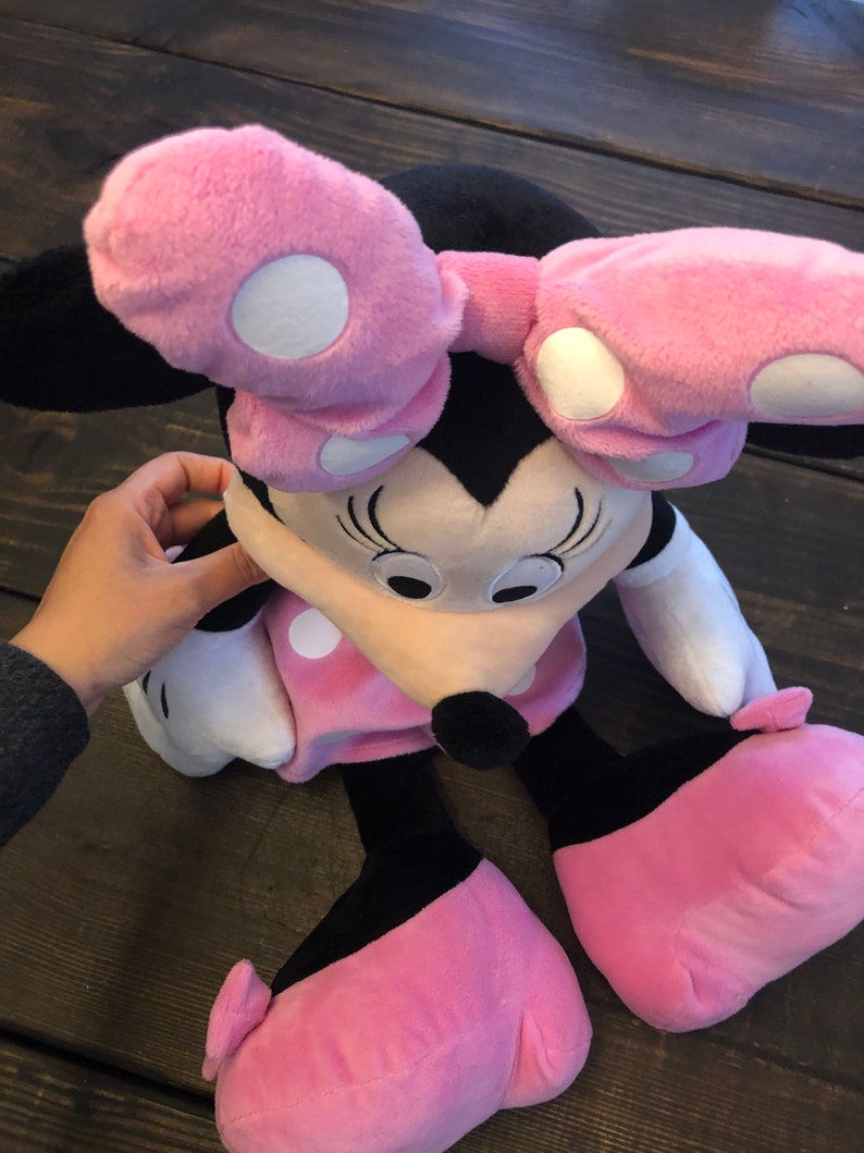 May include: Pink and black plush toy of Minnie Mouse with white polka dots. The toy has a large pink bow and is sitting on a wooden surface.