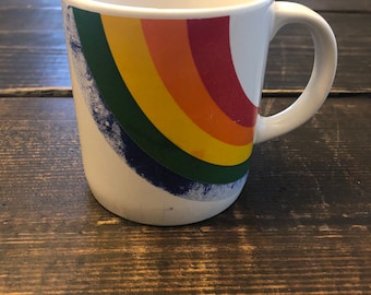 May include: White ceramic mug with a rainbow design. The rainbow is made up of red, orange, yellow, green, blue, and purple stripes. The mug has a blue and white speckled design on the bottom.