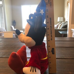 May include: A plush Mickey Mouse toy wearing a blue and white wizard hat and a red robe. The toy is sitting on a wooden surface and is about 17 inches tall.
