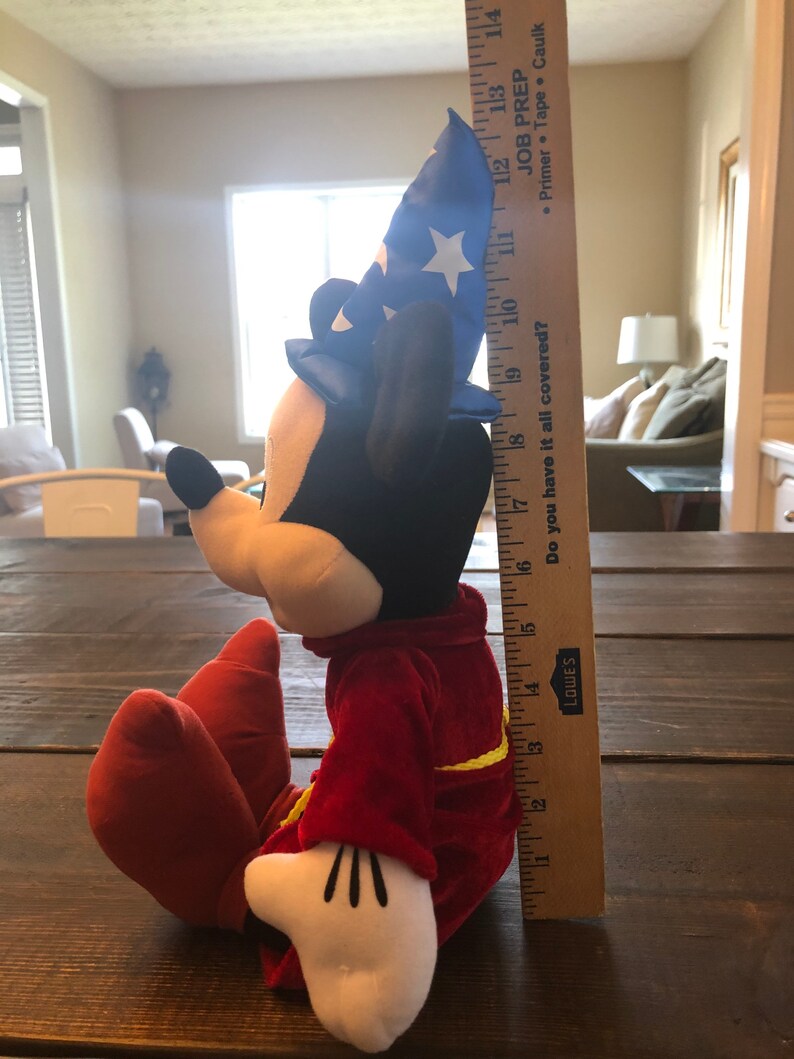 May include: A plush Mickey Mouse toy wearing a blue and white wizard hat and a red robe. The toy is sitting on a wooden surface and is about 17 inches tall.
