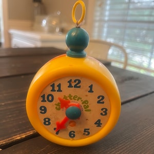 May include: A yellow, vintage, wind-up clock with a teal top and a red clock hand. The clock face has black numbers and the words "Sutter Pal" printed on it. The clock is sitting on a dark wooden surface.