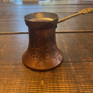 May include: A copper-colored, small coffee pot with a long, ornate handle. The pot has a flared base and a narrow top. The word "MOSTAR" is etched into the side, along with decorative patterns. The pot is sitting on a wooden table.