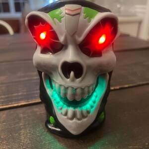 May include: A skull-shaped decorative light with glowing red eyes and a green illuminated mouth. The skull is white with black and green accents. The light is sitting on a wooden surface.