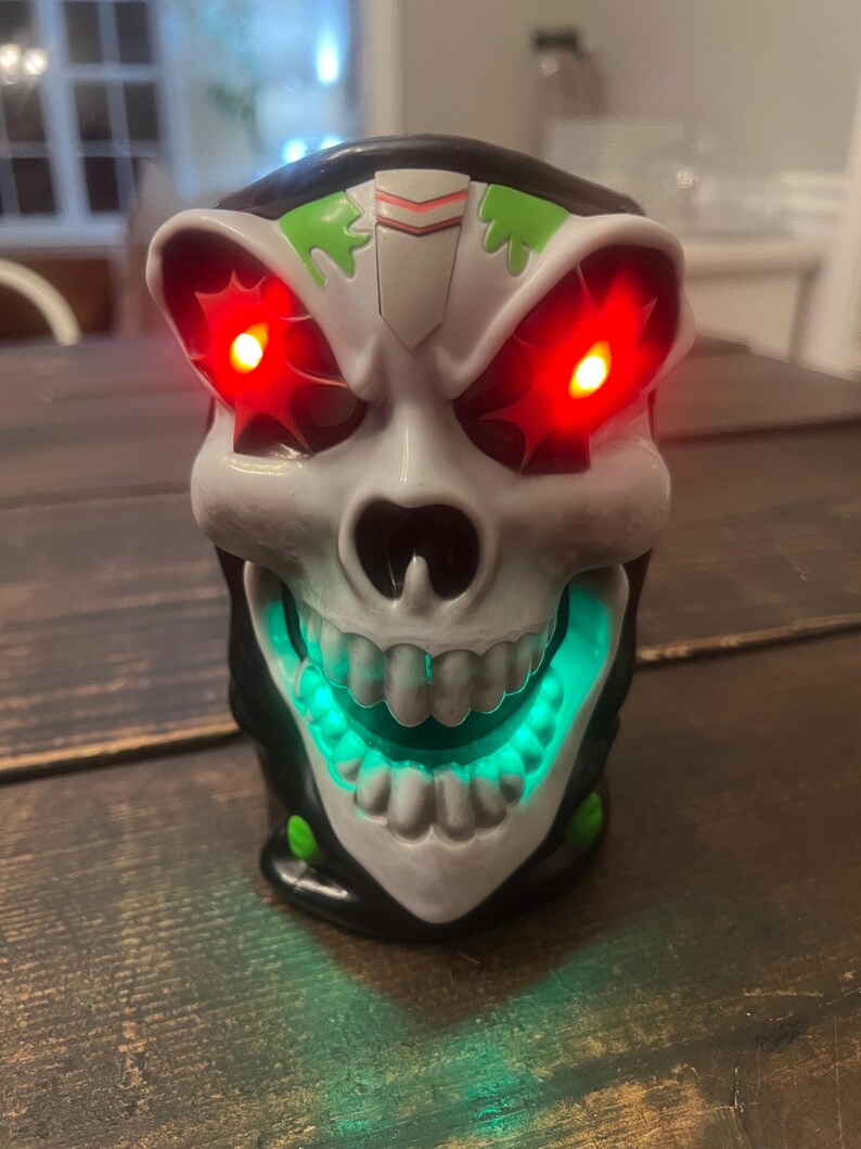 May include: A skull-shaped decorative light with glowing red eyes and a green illuminated mouth. The skull is white with black and green accents. The light is sitting on a wooden surface.