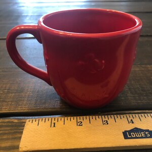 May include: A bright red ceramic mug with a handle. The mug is sitting on a wooden surface next to a ruler with the word "LOWE'S" printed on it.