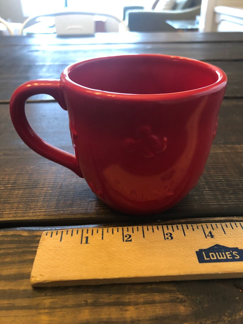 May include: A bright red ceramic mug with a handle. The mug is sitting on a wooden surface next to a ruler with the word "LOWE'S" printed on it.