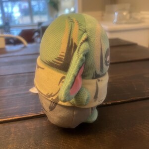 Pode incluir: Um brinquedo de pel&uacute;cia que retrata um personagem com orelhas grandes, cabe&ccedil;a verde e roupa bege e cinza. O brinquedo tem uma cabe&ccedil;a estampada em verde claro e bege com orelhas internas rosa. O brinquedo est&aacute; em uma superf&iacute;cie de madeira marrom escura.