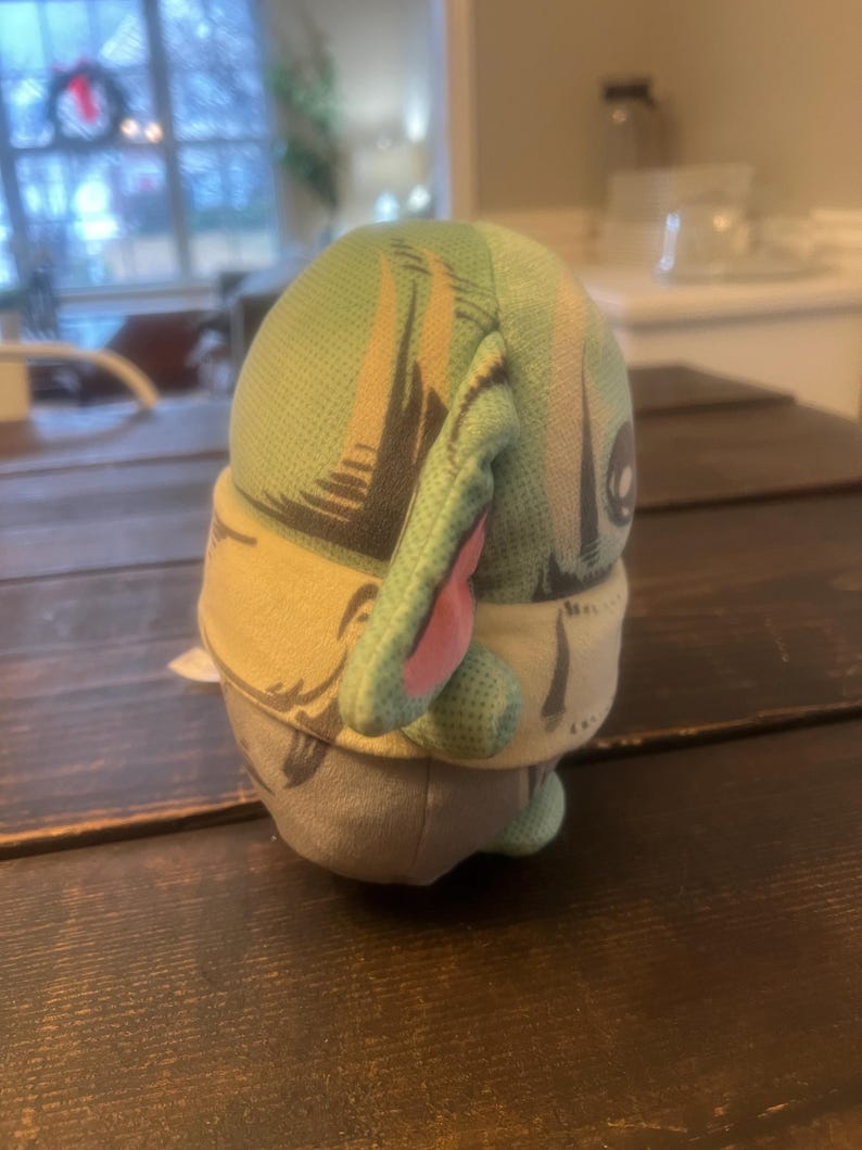 Pode incluir: Um brinquedo de pel&uacute;cia que retrata um personagem com orelhas grandes, cabe&ccedil;a verde e roupa bege e cinza. O brinquedo tem uma cabe&ccedil;a estampada em verde claro e bege com orelhas internas rosa. O brinquedo est&aacute; em uma superf&iacute;cie de madeira marrom escura.