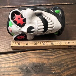 May include: A ceramic skull-shaped mug with a black top and green accents. The skull has white teeth and a white face with red starburst designs in the eye sockets. A wooden ruler is placed next to the mug.