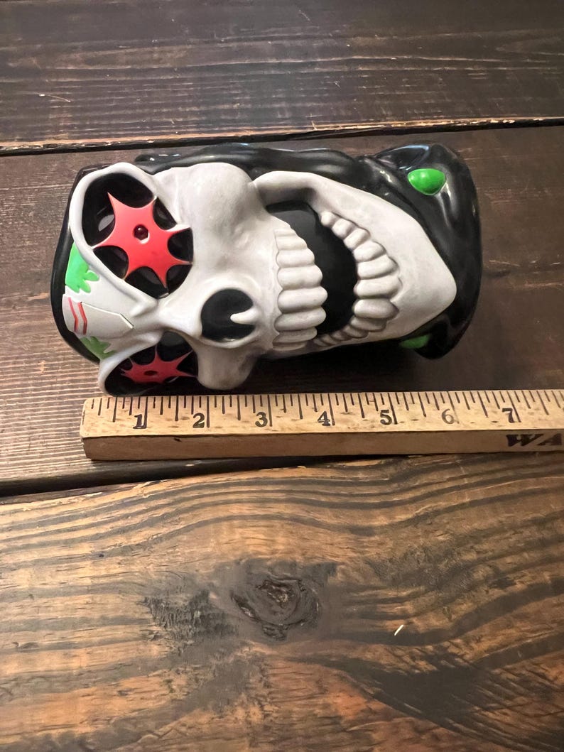 May include: A ceramic skull-shaped mug with a black top and green accents. The skull has white teeth and a white face with red starburst designs in the eye sockets. A wooden ruler is placed next to the mug.