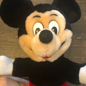 May include: A plush Mickey Mouse toy with black ears, a black body, red shorts with yellow buttons, and a white face with black eyes and a black nose.