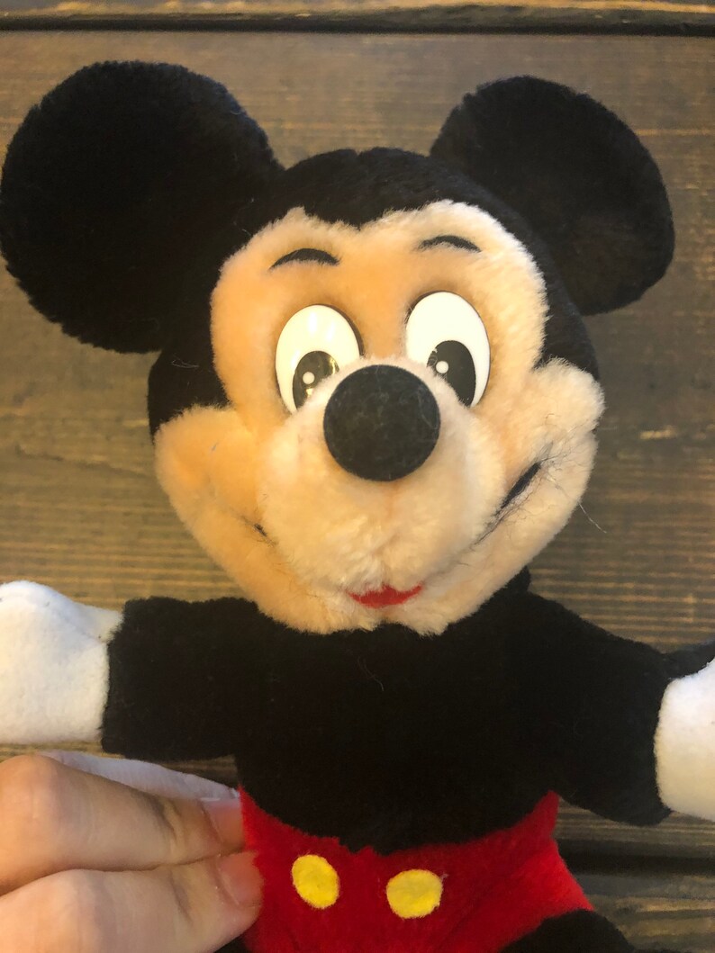 May include: A plush Mickey Mouse toy with black ears, a black body, red shorts with yellow buttons, and a white face with black eyes and a black nose.