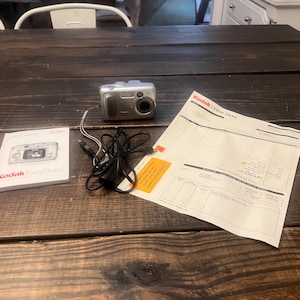 May include: A silver Kodak digital camera with a lens and flash is on a dark wooden table. A Kodak EasyShare manual, a power cord, and a receipt are also on the table.