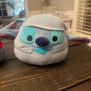 May include: A plush toy of a blue alien character with large turquoise eyes, a dark blue nose, and a light blue hat. The toy has large ears with pink accents and is sitting on a dark wooden surface.