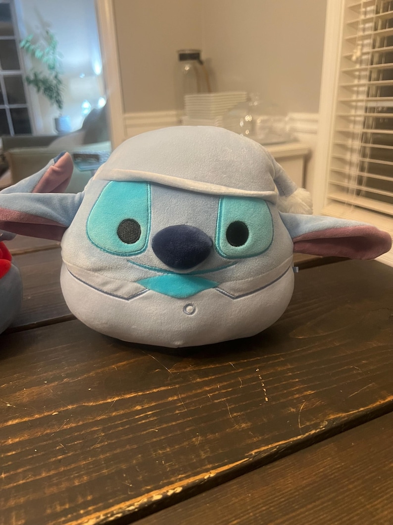 May include: A plush toy of a blue alien character with large turquoise eyes, a dark blue nose, and a light blue hat. The toy has large ears with pink accents and is sitting on a dark wooden surface.