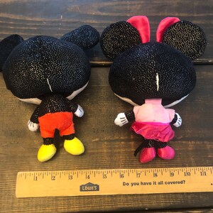 May include: A black and red plush Mickey Mouse and a black and pink plush Minnie Mouse. Both characters have sparkly black fabric and are standing on a wooden surface with a ruler.