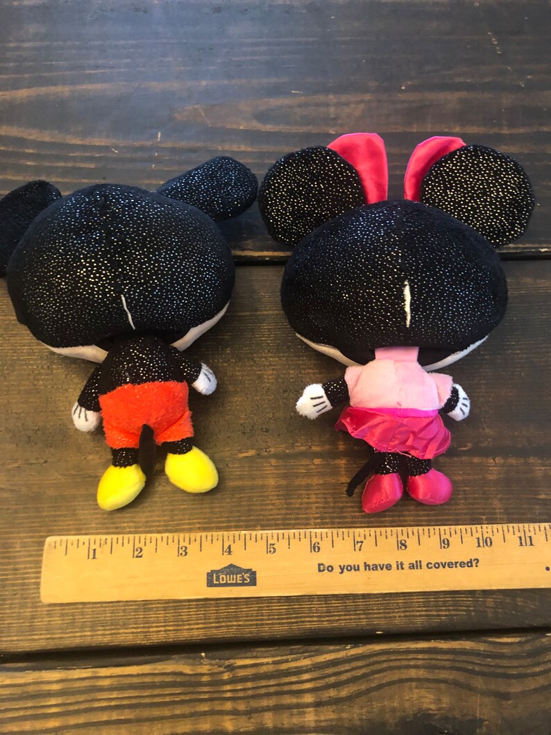 May include: A black and red plush Mickey Mouse and a black and pink plush Minnie Mouse. Both characters have sparkly black fabric and are standing on a wooden surface with a ruler.