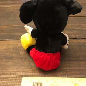 Puede incluir: Juguete de peluche Mickey Mouse negro y amarillo con pantalones cortos rojos sentado en una superficie de madera.