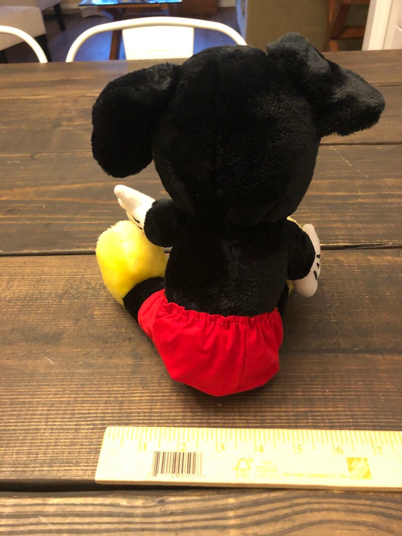 Puede incluir: Juguete de peluche Mickey Mouse negro y amarillo con pantalones cortos rojos sentado en una superficie de madera.