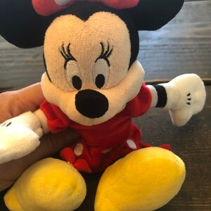 Op de afbeelding: Een pluche speelgoed van Minnie Mouse die een rode jurk draagt met witte stippen en een rode strik met witte stippen. Ze heeft zwarte oren, een zwarte neus en gele schoenen.