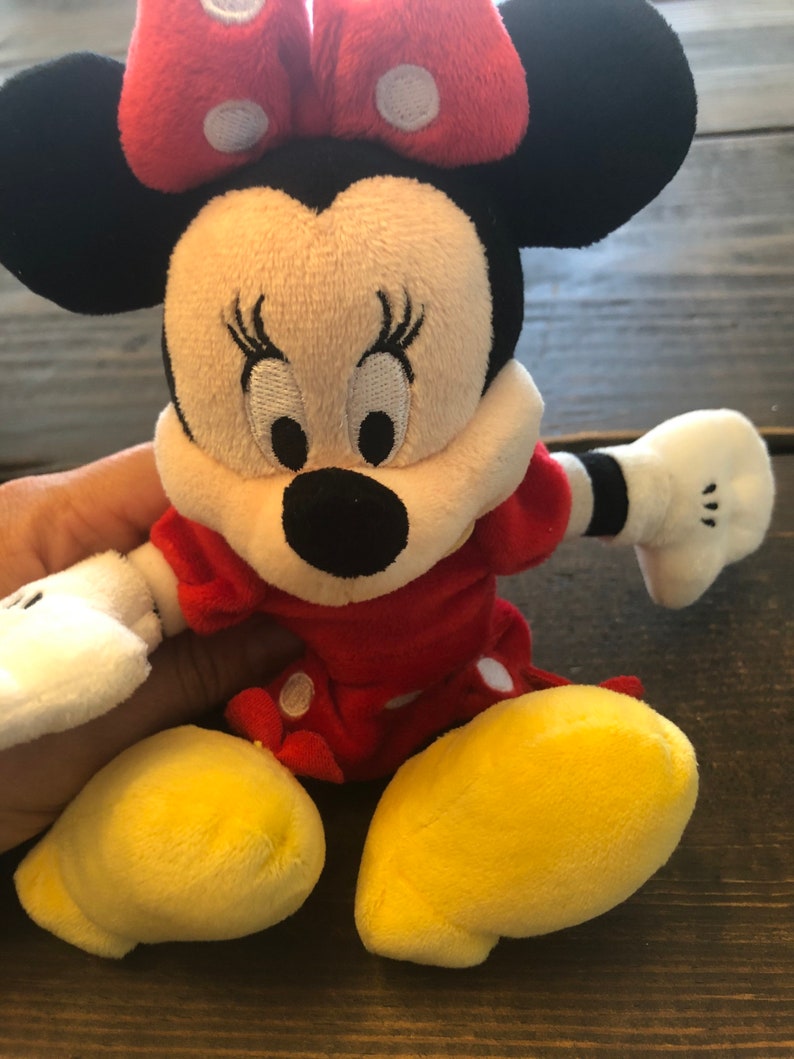 Op de afbeelding: Een pluche speelgoed van Minnie Mouse die een rode jurk draagt met witte stippen en een rode strik met witte stippen. Ze heeft zwarte oren, een zwarte neus en gele schoenen.