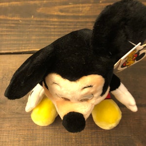 May include: A black and white plush Mickey Mouse toy with yellow shoes and a red shirt. The toy is sitting on a wooden surface.