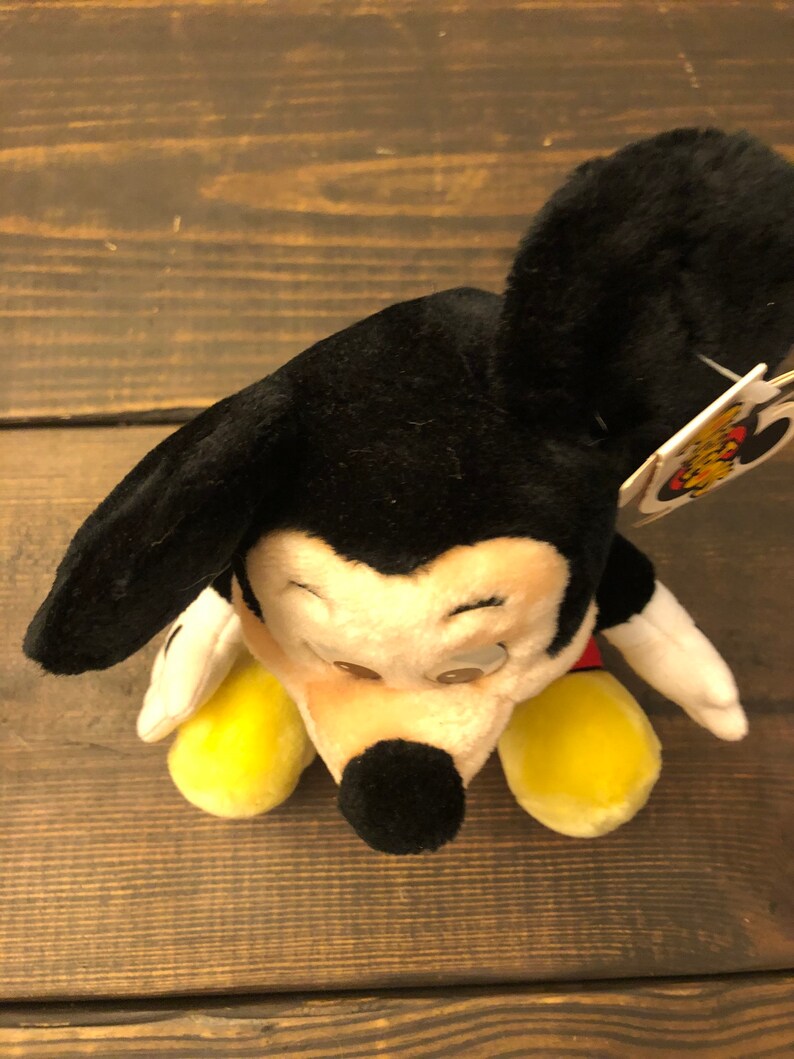 May include: A black and white plush Mickey Mouse toy with yellow shoes and a red shirt. The toy is sitting on a wooden surface.