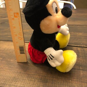 Puede incluir: Un juguete de peluche de Mickey Mouse sentado en una superficie de madera. El juguete es negro, rojo y amarillo. Lleva una camisa roja y blanca y zapatos amarillos.