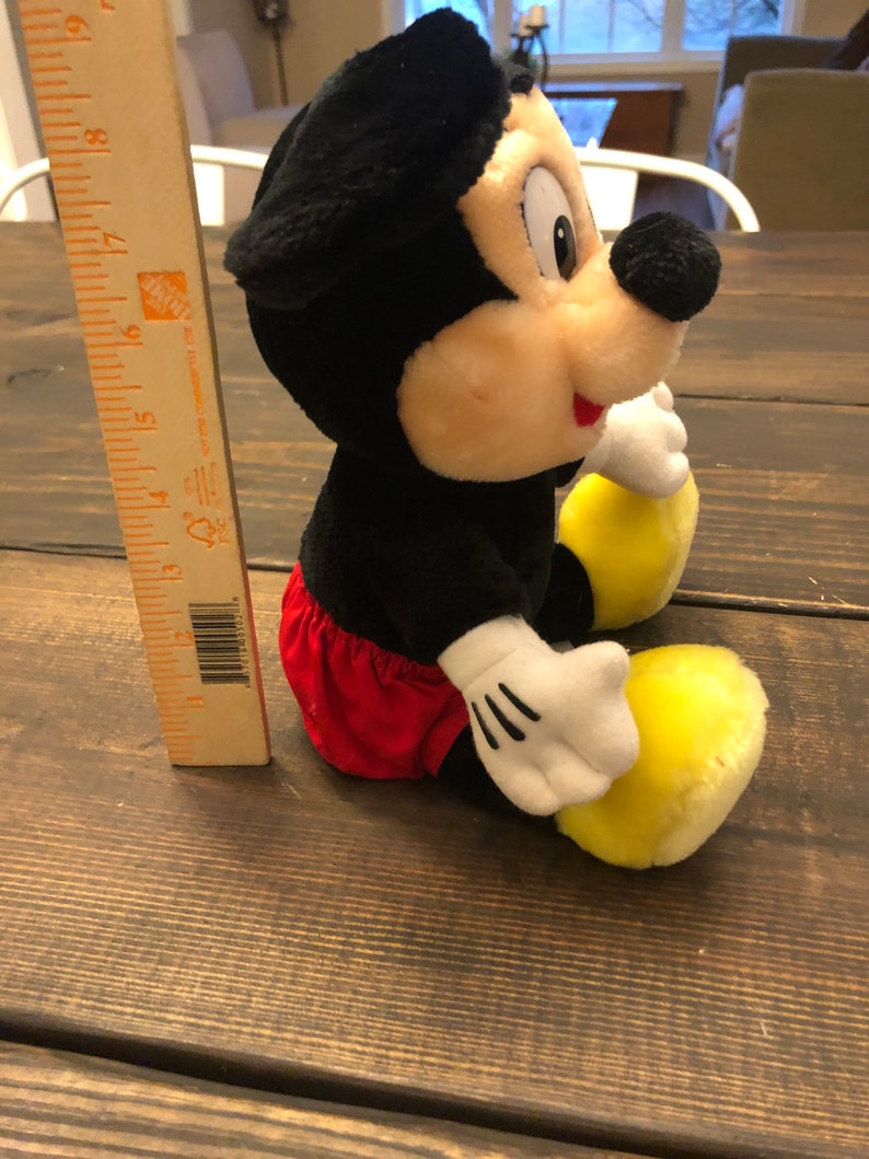 Puede incluir: Un juguete de peluche de Mickey Mouse sentado en una superficie de madera. El juguete es negro, rojo y amarillo. Lleva una camisa roja y blanca y zapatos amarillos.