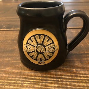 May include: Black ceramic mug with a gold circular design featuring the text "St. Kate's" and the word "DAD" below.