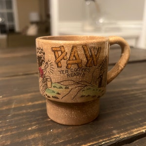 May include: A brown ceramic coffee mug with a cartoon illustration of a man holding a log and the text "Yer Coffee's Ready!!"