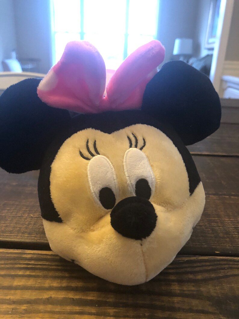May include: A plush toy of Minnie Mouse with a pink bow and black ears. The toy has a beige face with black eyes and a black nose.