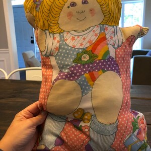 May include: A vintage Cabbage Patch Kids pillow featuring a girl with blonde hair wearing a purple and white polka dot outfit with a rainbow and stars design.