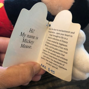 May include: A white tag with black text attached to a black and white Mickey Mouse plush toy. The tag reads "Hi! My name is Mickey Mouse." and "Open the door to enchantment and imagination...open your heart to magic brought to life in a playful collection of Disney friends created by Applause. Childhood dreams of fun with Mickey and his pals come true with plush playmates that capture the warmth and whimsy of Disney's best-loved characters and reflect the standards of quality that make every Applause product special. AM6 8-95"