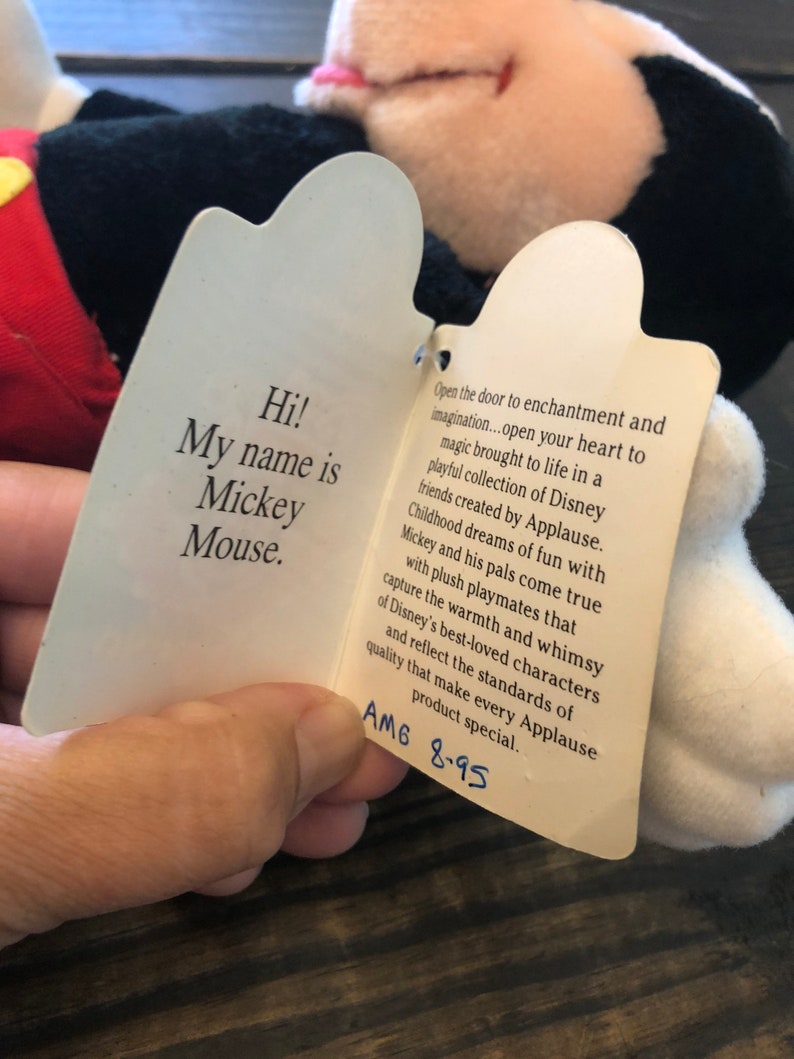 May include: A white tag with black text attached to a black and white Mickey Mouse plush toy. The tag reads "Hi! My name is Mickey Mouse." and "Open the door to enchantment and imagination...open your heart to magic brought to life in a playful collection of Disney friends created by Applause. Childhood dreams of fun with Mickey and his pals come true with plush playmates that capture the warmth and whimsy of Disney's best-loved characters and reflect the standards of quality that make every Applause product special. AM6 8-95"