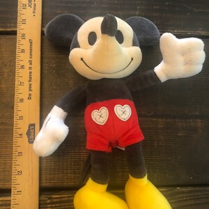 Puede incluir: Un juguete de peluche de Mickey Mouse con un cuerpo negro y gris, pantalones cortos rojos con dos corazones blancos y zapatos amarillos. El juguete sonr&iacute;e y tiene un guante blanco en su mano derecha.