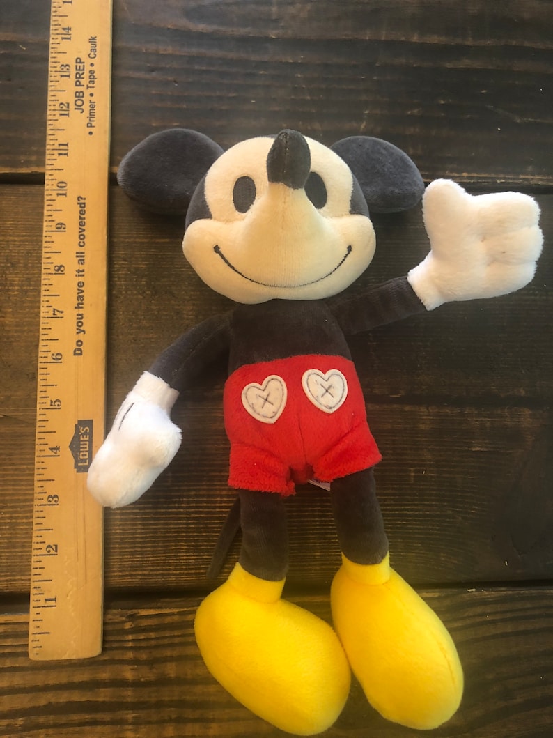 Puede incluir: Un juguete de peluche de Mickey Mouse con un cuerpo negro y gris, pantalones cortos rojos con dos corazones blancos y zapatos amarillos. El juguete sonr&iacute;e y tiene un guante blanco en su mano derecha.